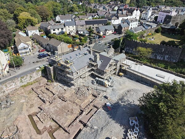 Archaeological excavation on the open ground beside Island House: cut trenches and exposed stone wall footings, with the house and the castle wall behind.