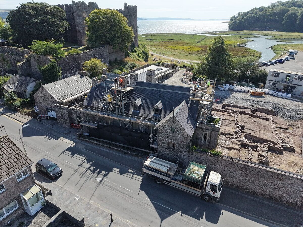 Island House close to, from the air: the scaffolded house below the castle wall, with the Taf estuary and the sea catching the light beyond.