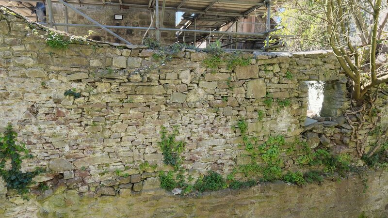 The creek-side wall of Island House today: a long rubble-stone boundary wall with blocked window openings, rising from the overgrown bed of the stream.